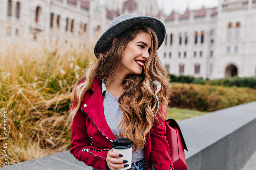 Fototapeta premium Joyful long-haired woman with trendy black manicure looking away while waiting someone near old building. Photo of pretty caucasian female model in hat drinking coffee outdoor.