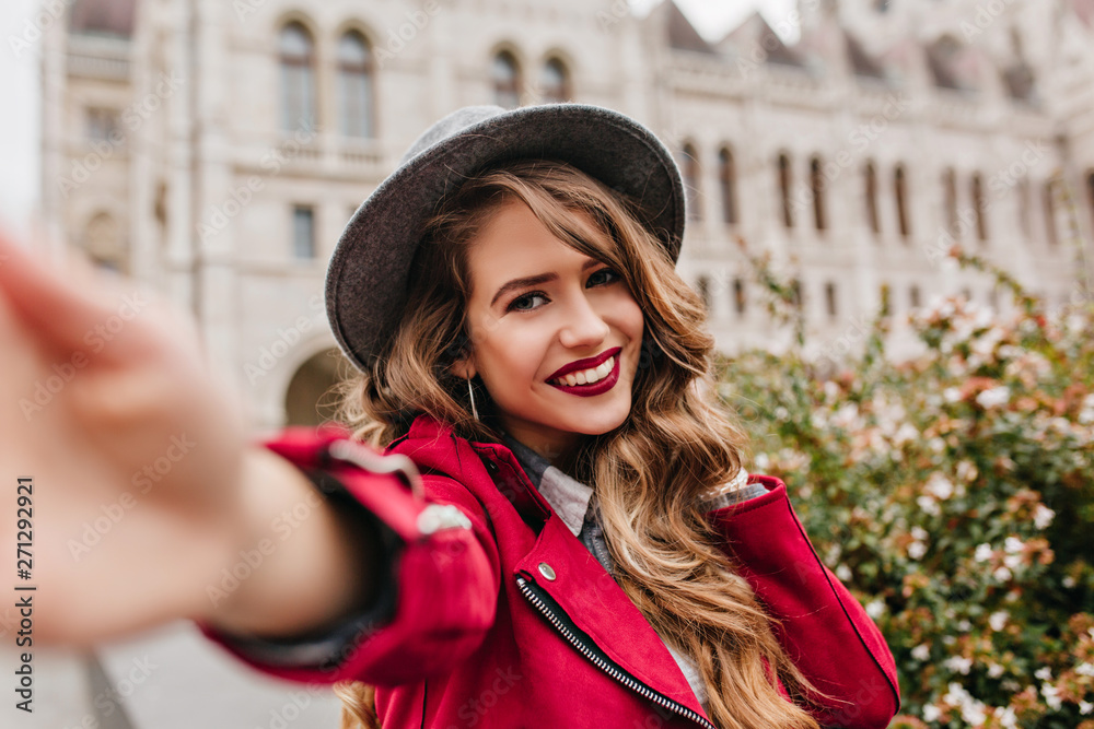 Joyful caucasian girl spending vacation in Hungary and making selfie. Outdoor portrait of funny elegant lady in black hat taking picture of herself, walking past old building.