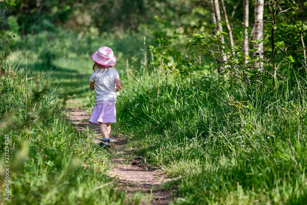 Rear view of young 3 year old girl with water bottle in her hand walking on a narrow path in the green summer forest