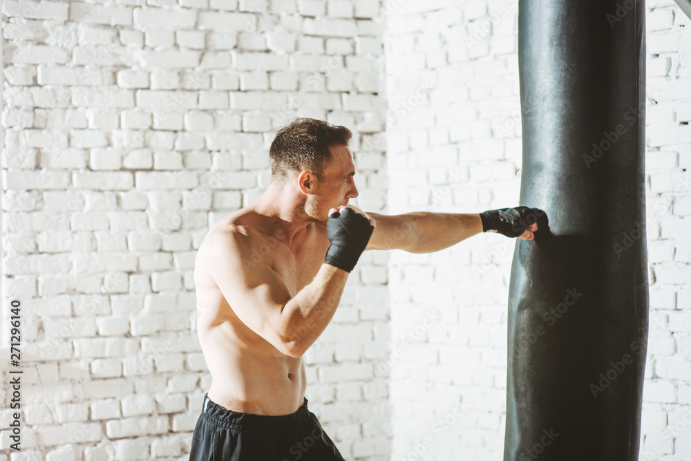 Muscular fighter practicing with punching bag against white brick wall.