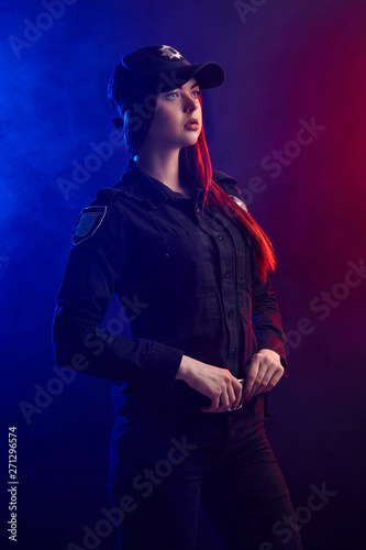 Serious female police officer is posing for the camera against a black background with red and blue backlighting. © nazarovsergey