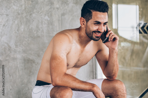 Hispanic man in shorts smiling and answering phone call while sitting on bench during training in gym