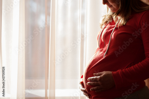 Beautiful young pregnant woman standing by the window at home. Expectant mother with a big belly