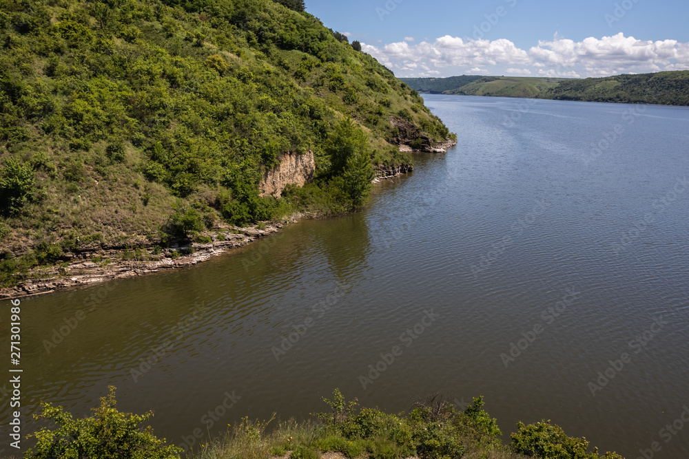 The high and steep shore of the Dniester, covered with green grass and bushes. Clear sky and clouds on the horizon. Summer landscape. Ukraine.