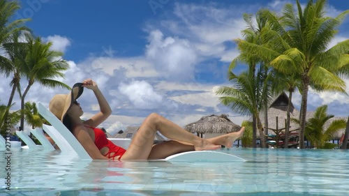 Travel luxury hotel swimming pool woman relaxing in lounging chair enjoying summer vacation. Tourist with hat and red swimsuit in water lounger.