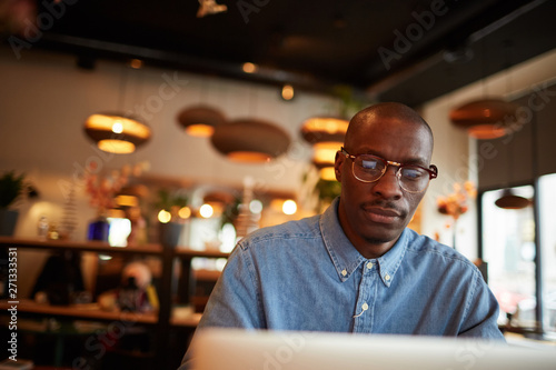 Warm toned portrait of contemporary African-American man using laptop sitting at table in cafe, copy space
