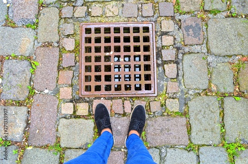 feet standing in front of the square grille of the manhole on the background of a paved road © Anastassiya