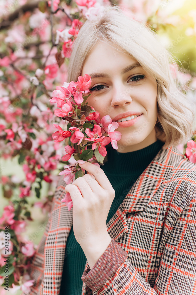 Fototapeta premium Close up portrait of young pretty caucasian curly blondie model posing outdoors in pink blossom garden. Beauty, nature concept