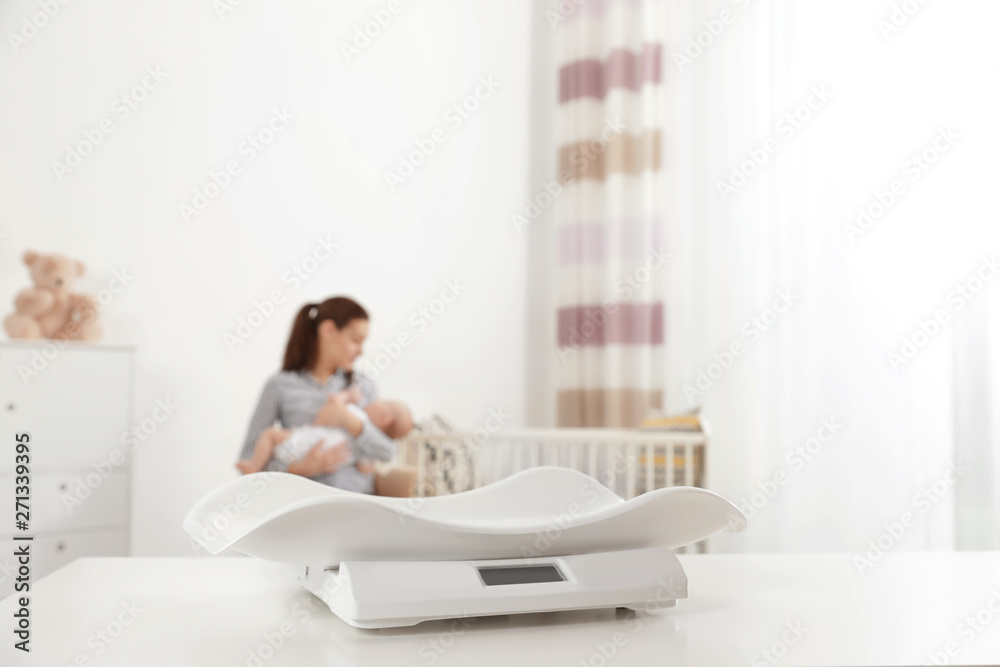 Modern baby scales on table and blurred woman with child indoors. Space for text Modern baby scales on table and blurred woman with child indoors. Space for text