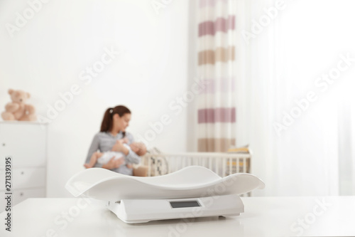 Modern baby scales on table and blurred woman with child indoors. Space for text