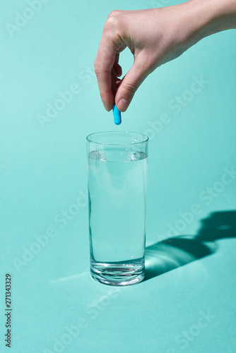 cropped view of woman putting blue pill in transparent glass with fresh water on turquoise background