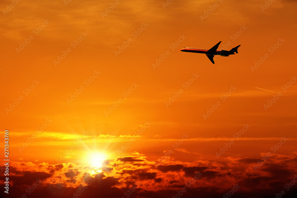 Silhouetted flying airplane over beautiful cloudscape of orange colored sunset sky with shining sun