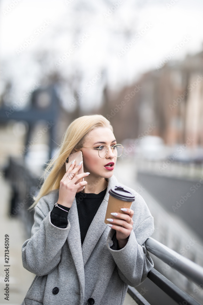 Cheerful young woman wearing coat talk her phone in the sunny city street and drinking take away coffee in paper cup. Cheerful young woman wearing coat talk her phone in the sunny city street and drinking take away coffee in paper cup.