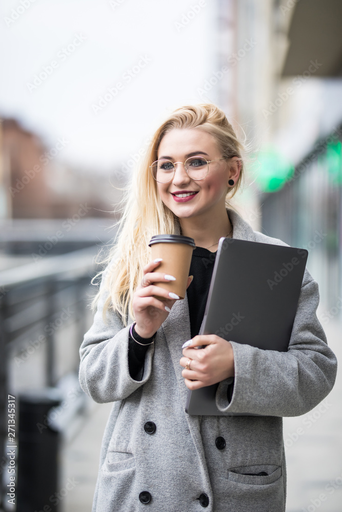 Portrait of smiling woman strolling through city street with silver laptop and takeaway coffee in hands Portrait of smiling woman strolling through city street with silver laptop and takeaway coffee in hands