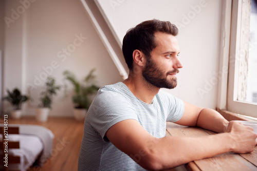 Young Man Relaxing In Loft Apartment Looking Out Of Window With Hot Drink
