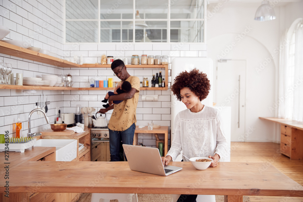 Multi-Tasking Father Holds Baby Son And Pours Drink As Mother Uses Laptop And Eats Breakfast Multi-Tasking Father Holds Baby Son And Pours Drink As Mother Uses Laptop And Eats Breakfast