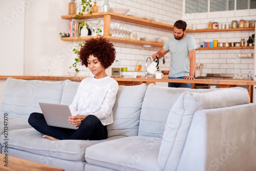 Woman Relaxing Sitting On Sofa At Home Using Laptop Computer With Man In Kitchen Pouring Drink