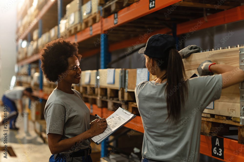 Happy female African American dispatcher and worker communicating in distribution warehouse.