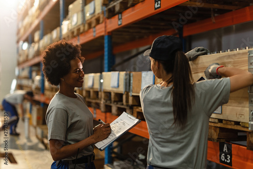 Happy female African American dispatcher and worker communicating in distribution warehouse.