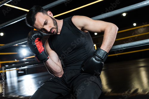 low angle view of tired bearded man in black boxing gloves touching head in gym