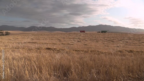 An end of day wide vista of a farm field during harvest with agricultural machinery in the background and mountains and clouds as a backdrop.