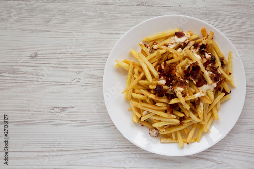 French fries with cheese sauce and bacon on a white plate over white wooden surface, top view. Flat lay, from above, overhead. Copy space.