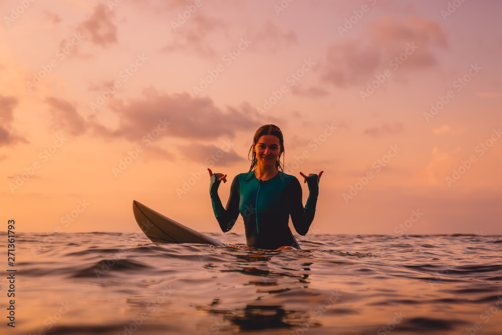 Smiling surfer woman sit on a surfboard in ocean. Surfing at sunset
