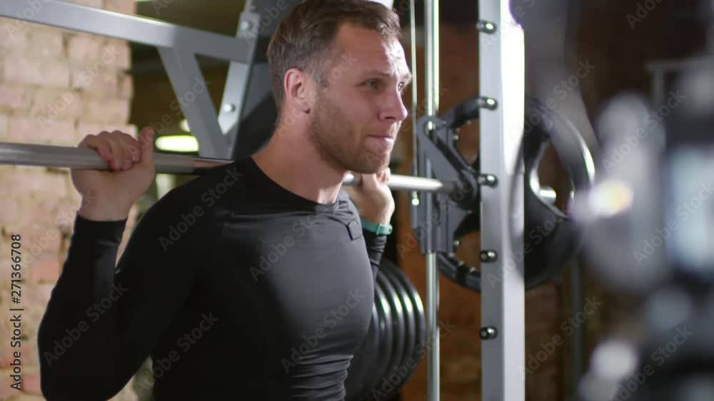 Rack focused shot of male bodybuilder saying something on camera and then doing barbell squats in gym while recording video lesson for sports channel Rack focused shot of male bodybuilder saying something on camera and then doing barbell squats in gym while recording video lesson for sports channel