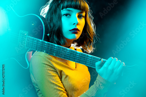 Beautiful young hipster woman with curly hair with red guitar in neon lights. Rock musician is playing electrical guitar. 90s style concept.