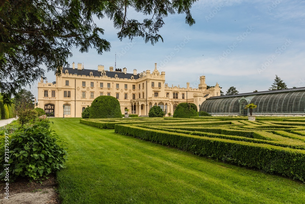 Obraz premium Lednice, Czech Republic - May 28 2019: Famous Lednice castle in South Moravia with yellow facade. Garden with green lawn, bushes, trees and greenhouse. Sunny spring day, blue sky, white clouds.