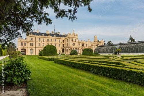 Lednice, Czech Republic - May 28 2019: Famous Lednice castle in South Moravia with yellow facade. Garden with green lawn, bushes, trees and greenhouse. Sunny spring day, blue sky, white clouds.