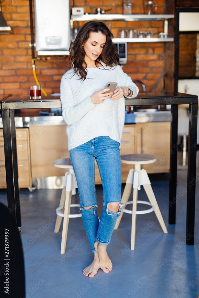 Young pretty woman using smartphone leaning at kitchen table in a modern home. Smiling woman reading phone message. Brunette happy girl typing a text message