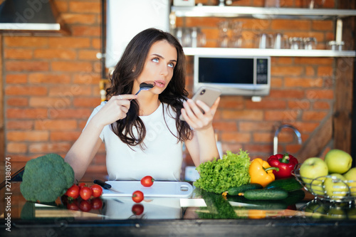 Young happy woman holding spoon and choose recipe with eco vegetables, look into web the phone and sitting in the kitchen © F8  \ Suport Ukraine