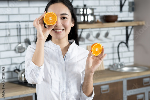 Front view of asian woman covering her one eye with orange slice in kitchen