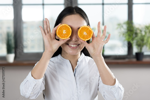 Smiling woman holding slices of orange in front of her eyes at home
