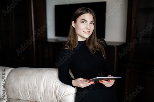Beautiful young woman in casual clothes is using a digital tablet and smiling while sitting on couch at home
