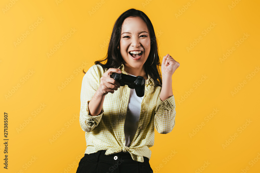 Obraz premium KYIV, UKRAINE - APRIL 16, 2019: happy asian girl playing video game with joystick, isolated on yellow