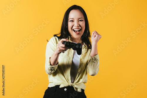 KYIV, UKRAINE - APRIL 16, 2019: happy asian girl playing video game with joystick, isolated on yellow