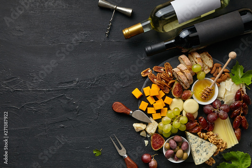 Top view of tasty cheese plate and wine bottles with fruit, grape, nuts and honey on a circle kitchen plate on the black stone background, top view, copy space. Gourmet food and drink.