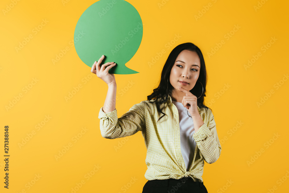 thoughtful asian woman holding green speech bubble, isolated on yellow thoughtful asian woman holding green speech bubble, isolated on yellow
