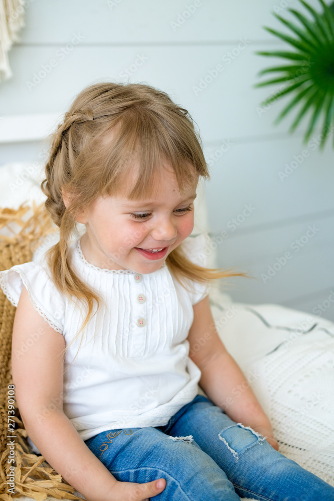 Beautiful little girl sitting on the bed in the bedroom. the baby has blonde hair and blue eyes. Jeans and white t-shirt Beautiful little girl sitting on the bed in the bedroom. the baby has blonde hair and blue eyes. Jeans and white t-shirt