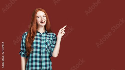 Look at it. Redhead smiling girl pointing with finger standing against red background