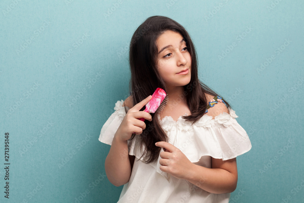 Portrait of healthy beautiful teenager brunette girl in white dress standing and combing brunette hair with pink hairbrush. Morning care. Indoor, isolated, studio shot, blue background Portrait of healthy beautiful teenager brunette girl in white dress standing and combing brunette hair with pink hairbrush. Morning care. Indoor, isolated, studio shot, blue background