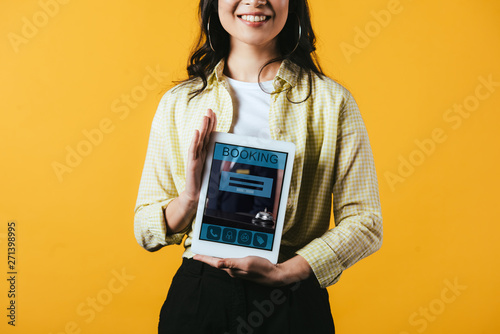 cropped view of smiling girl showing digital tablet with booking app, isolated on yellow