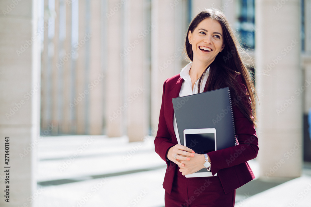 Portrait of business woman dressed in maroon suit, holds tablet and folder, smiling widely outdoor. Business people concept. Portrait of business woman dressed in maroon suit, holds tablet and folder, smiling widely outdoor. Business people concept.