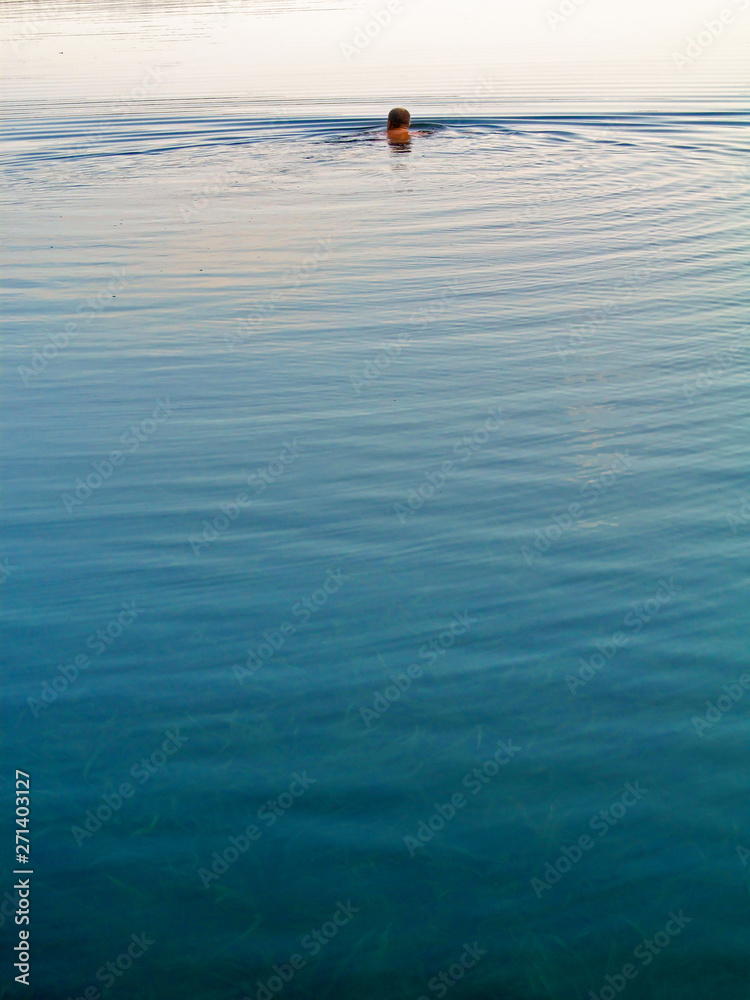 Swimmer swims away outdoor. Alone person moves in calm natural water, creating waves on a tranquil surface - back view. 