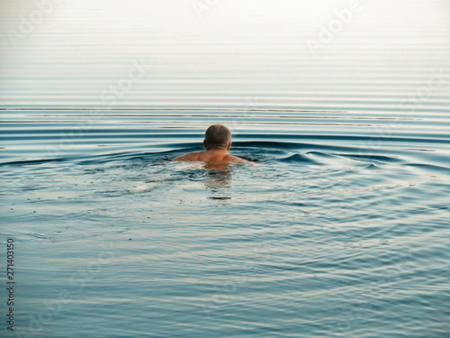 Swimmer swims away outdoor. Alone person moves in calm natural water  creating waves on a tranquil surface - back view. 