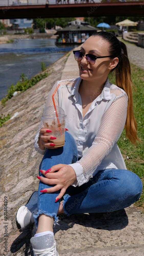 Young woman sitting by the river, drinking coffee and smiles