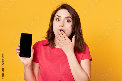 Image of attractive shocked young girl dressed red casual t shirt, has dark hair, covering mouth with hand holding smart phone with blank empty screen, looks at camera isolated over yellow background. photo