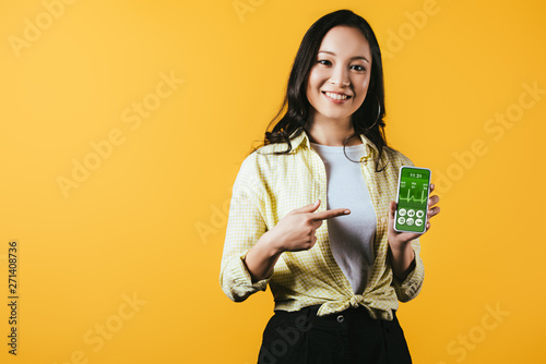 smiling asian girl pointing at smartphone with health app, isolated on yellow